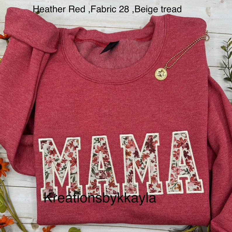 May include: Heather red sweatshirt with the word "MAMA" in large, floral-filled letters. A gold necklace with a pendant rests on the shirt. The text "Kreationsbykkayla" is at the bottom.