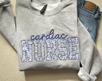 Op de afbeelding: Een lichtgrijs sweatshirt met ronde hals en de geborduurde tekst cardiac NURSE. Het woord cardiac is in blauw cursief, terwijl NURSE in blokletters is gevuld met een blauw-wit bloemenpatroon en omlijnd met blauwe draad.