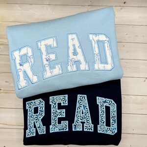 May include: Two sweatshirts, one pale blue and one navy, each displaying the word "READ" in large, decorative lettering. The letters on the pale blue sweatshirt have a floral pattern, while the navy sweatshirt's letters feature a daisy design.
