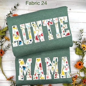 May include: Two folded green sweatshirts. The top one says "AUNTIE" in white block letters filled with a floral pattern. The bottom one says "MAMA" in the same style. The text "Fabric 24" is in the top left corner.
