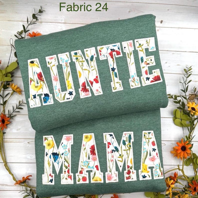 May include: Two folded green sweatshirts. The top one says "AUNTIE" in white block letters filled with a floral pattern. The bottom one says "MAMA" in the same style. The text "Fabric 24" is in the top left corner.