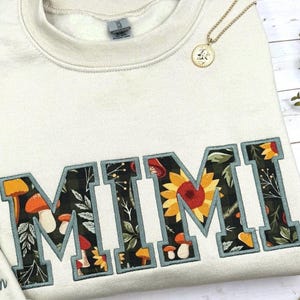 May include: Cream-colored sweatshirt with the embroidered word "MIMI" in a collegiate style. The letters are filled with a floral and mushroom pattern, outlined in teal. A gold pendant necklace rests on the sweatshirt. The text "Fabric 21 : Heather Green thread" is at the bottom.
