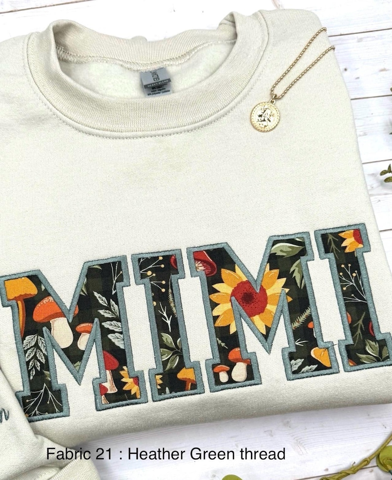 May include: Cream-colored sweatshirt with the embroidered word "MIMI" in a collegiate style. The letters are filled with a floral and mushroom pattern, outlined in teal. A gold pendant necklace rests on the sweatshirt. The text "Fabric 21 : Heather Green thread" is at the bottom.