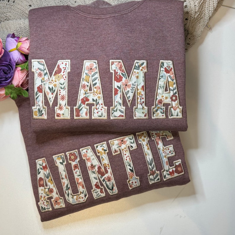 May include: Two mauve sweatshirts, one with "MAMA" and the other with "AUNTIE" in floral-patterned letters. The floral design includes red, yellow, and white flowers. The sweatshirts are folded and stacked on a white surface.