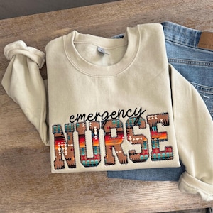 Op de afbeelding: Een beige sweater met ronde hals met het woord emergency in zwarte cursieve letters boven het woord NURSE in blokletters. De letters van NURSE zijn gevuld met een veelkleurig geometrisch patroon in tinten oranje, rood, blauw en bruin.