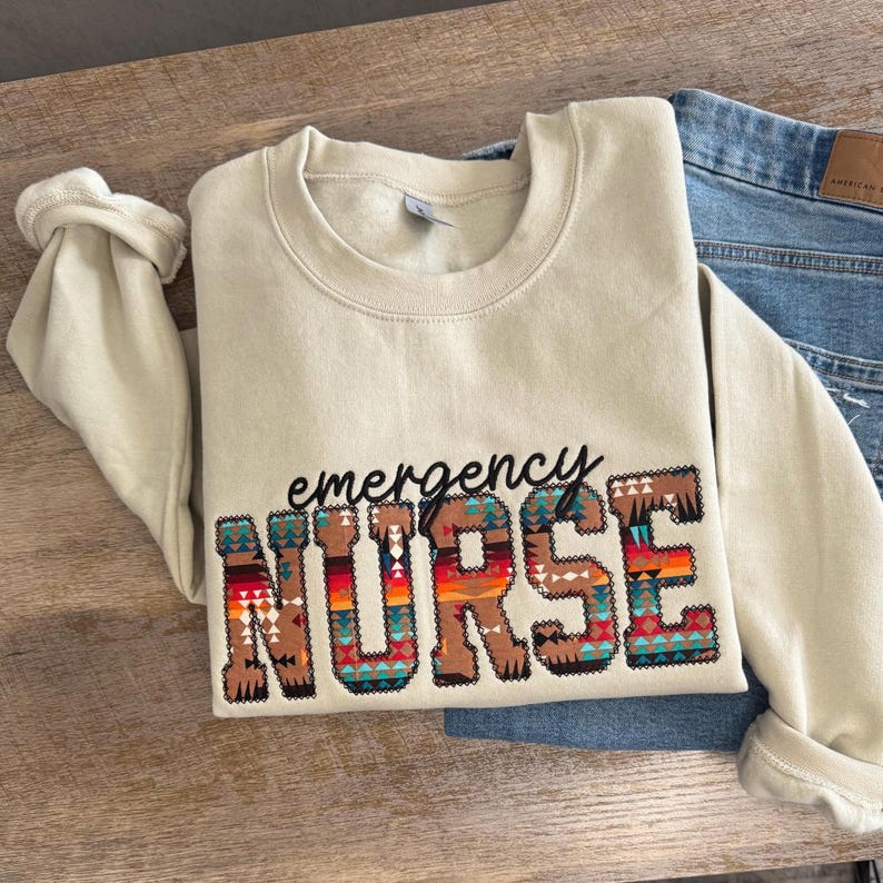 Op de afbeelding: Een beige sweater met ronde hals met het woord emergency in zwarte cursieve letters boven het woord NURSE in blokletters. De letters van NURSE zijn gevuld met een veelkleurig geometrisch patroon in tinten oranje, rood, blauw en bruin.