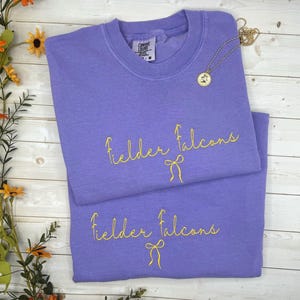 May include: Two lavender sweatshirts with the text "Fielder Falcons" in yellow script and a yellow bow graphic. A gold necklace with a pendant is draped across the top sweatshirt. The sweatshirts are folded on a light wood surface.