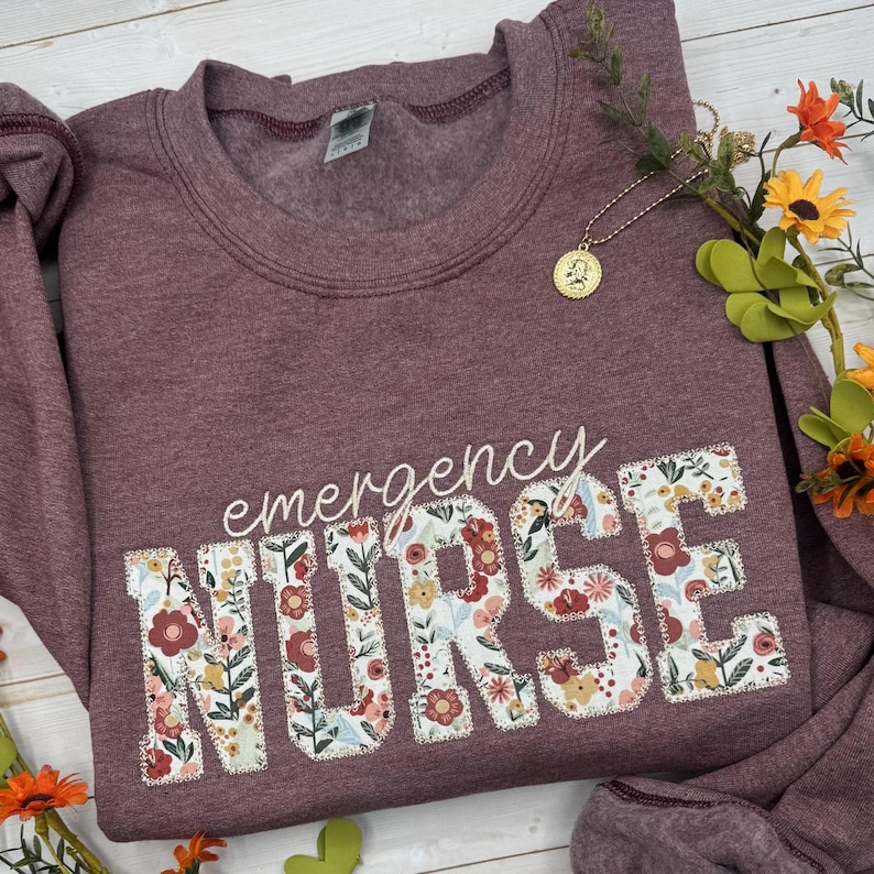 May include: Burgundy sweatshirt with the words "emergency NURSE" in white script and floral patchwork letters. A gold pendant necklace and flowers are arranged around the sweatshirt. The sweatshirt is made of a soft material.