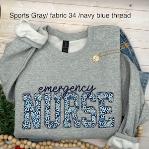 May include: A grey sweatshirt featuring the words "emergency NURSE" in navy blue, floral-patterned letters. The sweatshirt has white lining on the cuffs and collar. The text "Sports Gray/ fabric 34 /navy blue thread" is above the sweatshirt.