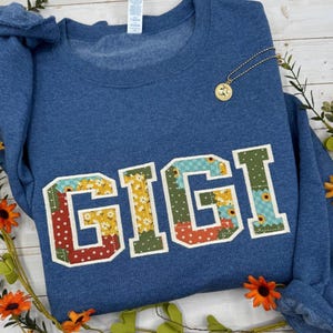 以下が含まれることがあります： パッチワーク風のカラフルな大きな文字で「GIGI」と書かれた青いスウェットシャツ。丸いペンダントが付いた金のネックレスがシャツの上にあります。スウェットシャツはオレンジ色の花と緑で飾られています。