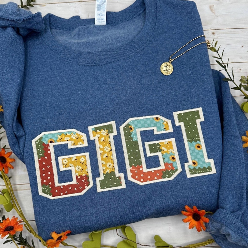 以下が含まれることがあります： パッチワーク風のカラフルな大きな文字で「GIGI」と書かれた青いスウェットシャツ。丸いペンダントが付いた金のネックレスがシャツの上にあります。スウェットシャツはオレンジ色の花と緑で飾られています。