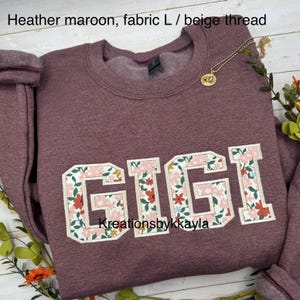 以下が含まれることがあります： 花柄の「GIGI」という名前が入った、ヘザーマルーンのスウェットシャツ。クルーネックと長袖です。「Kreationsbykkayla」の文字が下にあります。