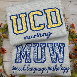 May include: A light gray sweatshirt featuring the blue-outlined yellow floral text UCD nursing and the blue-outlined white floral text MUW speech language pathology. The letters are appliqued with a bold block font style.