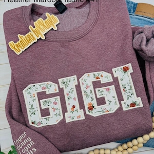 以下が含まれることがあります： フローラル文字で「GIGI」と書かれた、ヘザーマルーンのスウェットシャツ。スウェットシャツはクルーネックと長袖です。「Heather Maroon/ fabric #41」の文字が上にあります。袖には「Taylor Donny Logan Ellis」の名前があります。