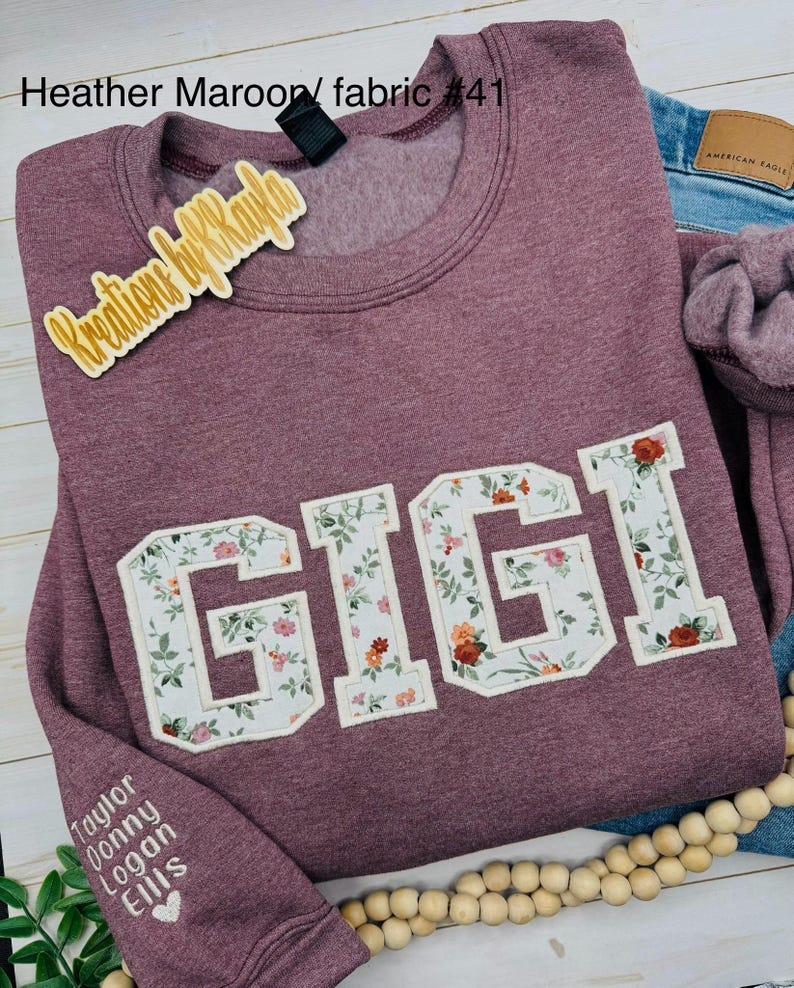 以下が含まれることがあります： フローラル文字で「GIGI」と書かれた、ヘザーマルーンのスウェットシャツ。スウェットシャツはクルーネックと長袖です。「Heather Maroon/ fabric #41」の文字が上にあります。袖には「Taylor Donny Logan Ellis」の名前があります。