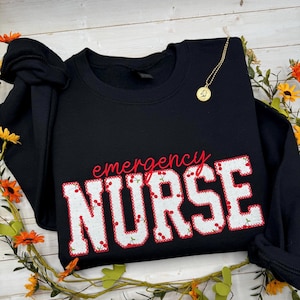 May include: Black sweatshirt featuring the text "emergency NURSE" in red, with a white and red cherry pattern. A gold necklace and floral decorations are also visible.
