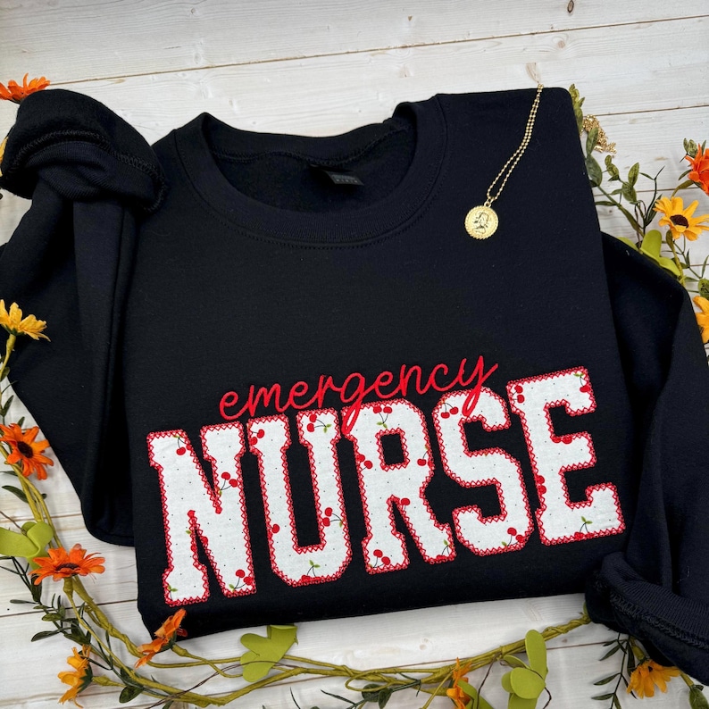 May include: Black sweatshirt featuring the text "emergency NURSE" in red, with a white and red cherry pattern. A gold necklace and floral decorations are also visible.