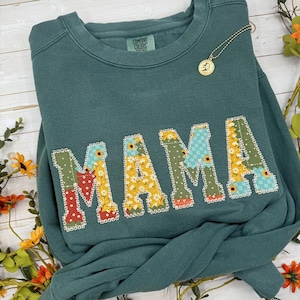 Puede incluir: Una sudadera Comfort Colors de color verde azulado con la palabra "MAMA" aplicada en la parte delantera. Las letras están decoradas con patrones florales y de patchwork. Un collar dorado con un colgante está sobre la sudadera.