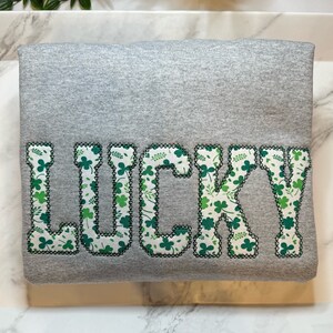 May include: Gray sweatshirt with the word "LUCKY" in large, green letters. The letters are outlined in black and filled with a pattern of green shamrocks.
