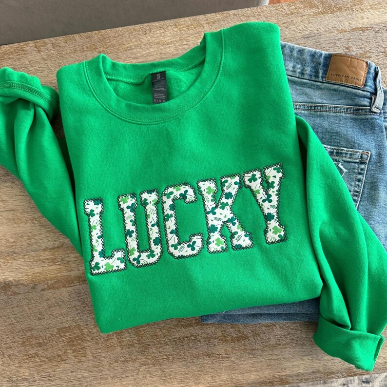 May include: Green sweatshirt with the word "LUCKY" in white letters outlined in black. The letters are filled with a pattern of green shamrocks.