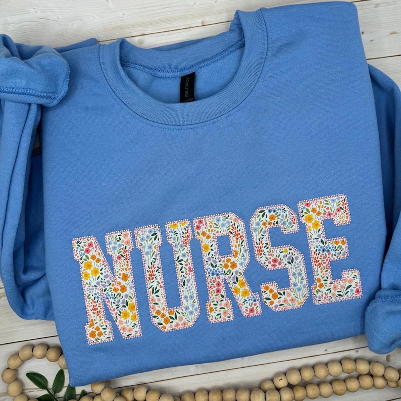 May include: A pale blue sweatshirt featuring the word "NURSE" in large, floral-patterned letters. The floral design incorporates a variety of colourful flowers. The sweatshirt has a crew neck and long sleeves.