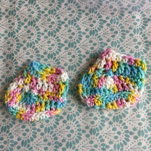Puede incluir: Dos pequeñas zapatillas de crochet en un patrón de colores arcoíris de rosa, amarillo, azul y blanco. Las zapatillas están sobre un fondo blanco con un patrón floral turquesa.