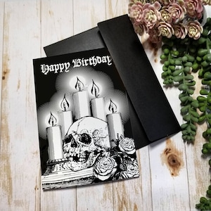 Black Greeting Card featuring a skull surrounded by glowing candles and roses. Gothic Script at the top of card reads "Happy Birthday". interior blank and white.