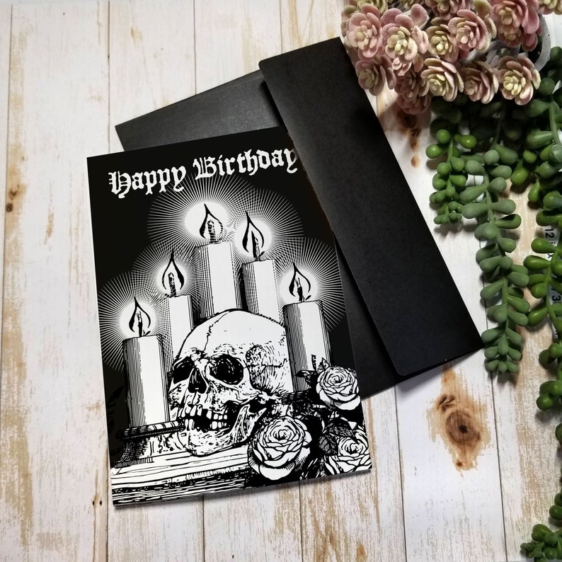 Black Greeting Card featuring a skull surrounded by glowing candles and roses. Gothic Script at the top of card reads "Happy Birthday". interior blank and white.