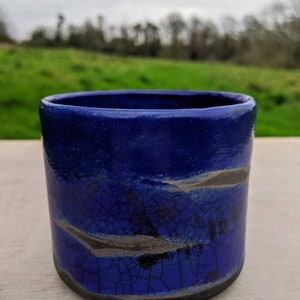 May include: A blue ceramic mug with a crackled glaze and black and silver accents. The mug is sitting on a wooden surface.