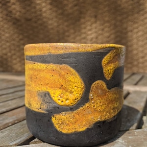 May include: A black ceramic mug with a yellow and orange glaze. The glaze has a textured, bubbly appearance.