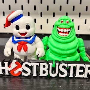 May include: A white Stay Puft Marshmallow Man figure with a blue and red sailor outfit and a green Slimer figure with a wide grin, positioned behind a black and white 3D-printed sign that reads GHOSTBUSTERS with a red ghost logo in the letter O.