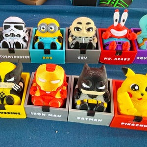 May include: A collection of thirteen small, colorful 3D-printed character figures, each seated in a matching square plastic box. The characters include Darth Vader, Stormtrooper, Bob, Gru, Mr. Krabs, Squidward, Plankton, Deadpool, Wolverine, Iron Man, Batman, Pikachu, and SpongeBob.