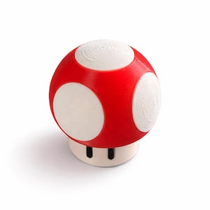 May include: A mushroom-shaped object with a bright red cap featuring circular white spots. The base is white and cylindrical, featuring two small black rectangular markings near the bottom. The surface texture shows concentric circular patterns.