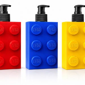 May include: Three rectangular soap dispensers shaped like large building blocks in red, blue, and yellow. Each dispenser features six raised circular studs on the front, each embossed with the LEGO logo, and a black plastic pump mechanism on top.