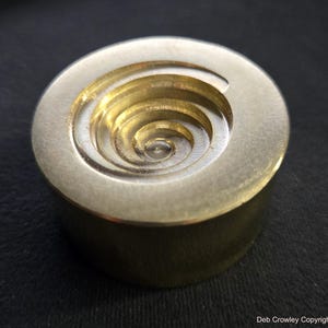 May include: A solid brass, circular object with a spiral design carved into the top. The spiral is recessed and appears to be a series of concentric rings. The object has a smooth, metallic finish and a cylindrical base.