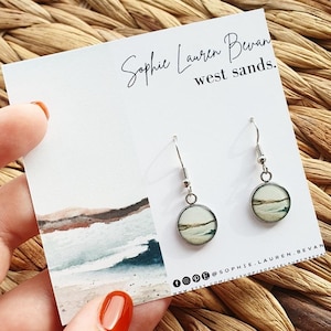 Può includere: Orecchini pendenti color argento con un design circolare a forma di paesaggio. Gli orecchini sono esposti su una carta bianca con la scritta "Sophie Lauren Bevan west sands". La carta presenta un acquerello di una scena di spiaggia. Gli orecchini sono tenuti da una mano.