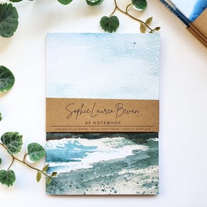 May include: An A5 notebook with a watercolour painting of a seascape on the cover. The cover is brown with the text "Sophie Lauren Bevan A5 Notebook 100% Recycled Paper 36 Unlined Pages Made in Scotland."