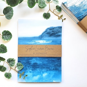 May include: A5 notebook with a blue seascape watercolour painting. The notebook is wrapped with a brown paper band that reads "Sophie Lauren Bevan" and "A5 Notebook". Made with 100% recycled paper, it has 36 unlined pages. Made in Scotland.