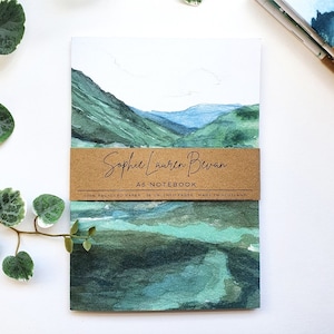 May include: A5 notebook with a watercolour landscape of green mountains and blue skies. The notebook has a brown band with the text "Sophie Lauren Bevan" and "A5 NOTEBOOK". The bottom of the band reads "100% RECYCLED PAPER 36 UNLINED PAGES MADE IN SCOTLAND."