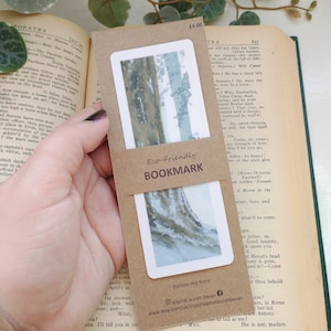 Può includere: Segnalibro ecologico con un dipinto ad acquerello di una scena boschiva con due alberi sullo sfondo. Il segnalibro è stampato su cartoncino marrone con il testo "Segnalibro ecologico" e "Seguimi qui" stampato su di esso. Il segnalibro include anche un indirizzo del sito web e un identificatore dei social media.