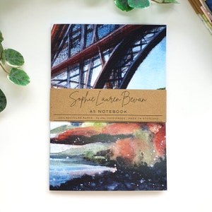 May include: A5 notebook with a watercolor painting of a bridge on the front cover. The notebook is made of 100% recycled paper and has 36 unlined pages. The notebook is made in Scotland.