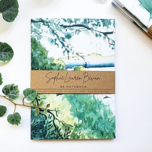 Puede incluir: Un cuaderno de acuarela con un diseño de paisaje verde y azul. La portada presenta el texto "Sophie Lauren Bevan A5 Notebook 100% Recycled Paper - 36 Unlined Pages - Made in Scotland".