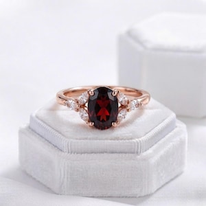 Op de afbeelding: Een ring van roségoud met een centrale, ovaal geslepen, dieprode granaatsteen. De ringband is aan weerszijden versierd met drie helderwitte, marquise-geslepen stenen, wat zorgt voor een symmetrische opstelling rondom de donkerrode steen.