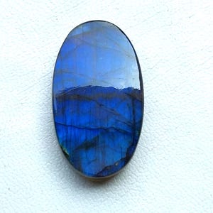 Puede incluir: Gema de labradorita de forma ovalada con superficie pulida. La piedra muestra un color azul vibrante con patrones oscuros e irregulares y una sutil iridiscencia. La gema está sobre un fondo blanco.