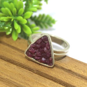 Puede incluir: Un anillo de plata con un engaste triangular lleno de pequeñas piedras preciosas de color rojo oscuro. El anillo descansa sobre una superficie de madera, con follaje verde en el fondo. La banda del anillo tiene un diseño de doble capa.