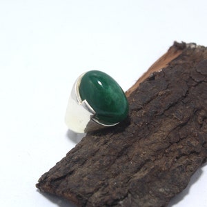 May include: A silver ring with a large, oval-shaped green gemstone set in the center. The ring is resting on a piece of rough-hewn wood.