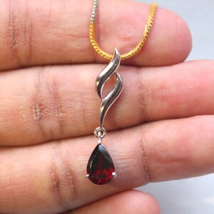 Pode incluir: Pingente de prata com uma pedra preciosa vermelha escura em forma de gota. O pingente tem um design em espiral e pende de uma corrente de ouro e prata. A pedra preciosa tem aproximadamente 1,3 cm de comprimento.
