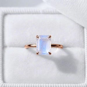May include: A ring featuring a rectangular emerald-cut moonstone set in a polished rose gold band. The gemstone displays a translucent, iridescent blue glow, held securely by four prongs on a thin, smooth metal band with a reflective finish.