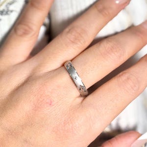 Könnte beinhalten: Ein silberner Ring mit floralem Muster und kleinen, runden Verzierungen. Der Ring wird an einem Finger getragen und zeigt sein aufwendiges Design und seine polierte Oberfläche. Das Band des Rings weist ein sich wiederholendes Muster aus Blättern und Punkten auf.