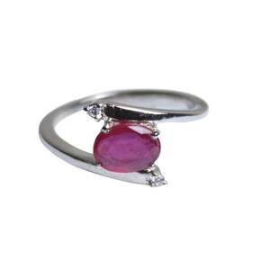 May include: A silver ring featuring a large, oval-cut ruby gemstone as the focal point. The ring has a modern, asymmetrical design with a curved band and small accent stones on either side of the ruby. The ruby is a deep red color.